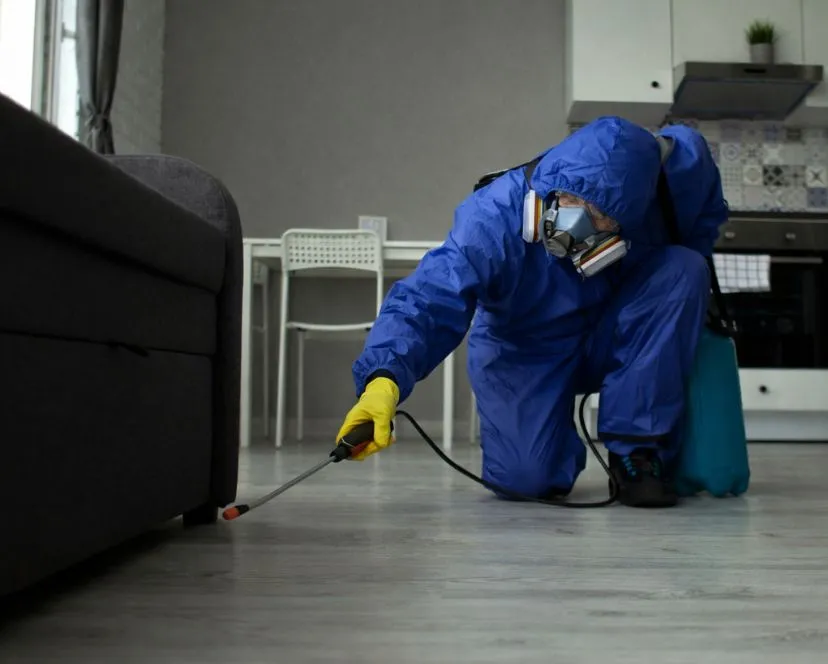 Professional exterminator in a blue safety suit and respirator spraying for pests in a large industrial warehouse storage area.